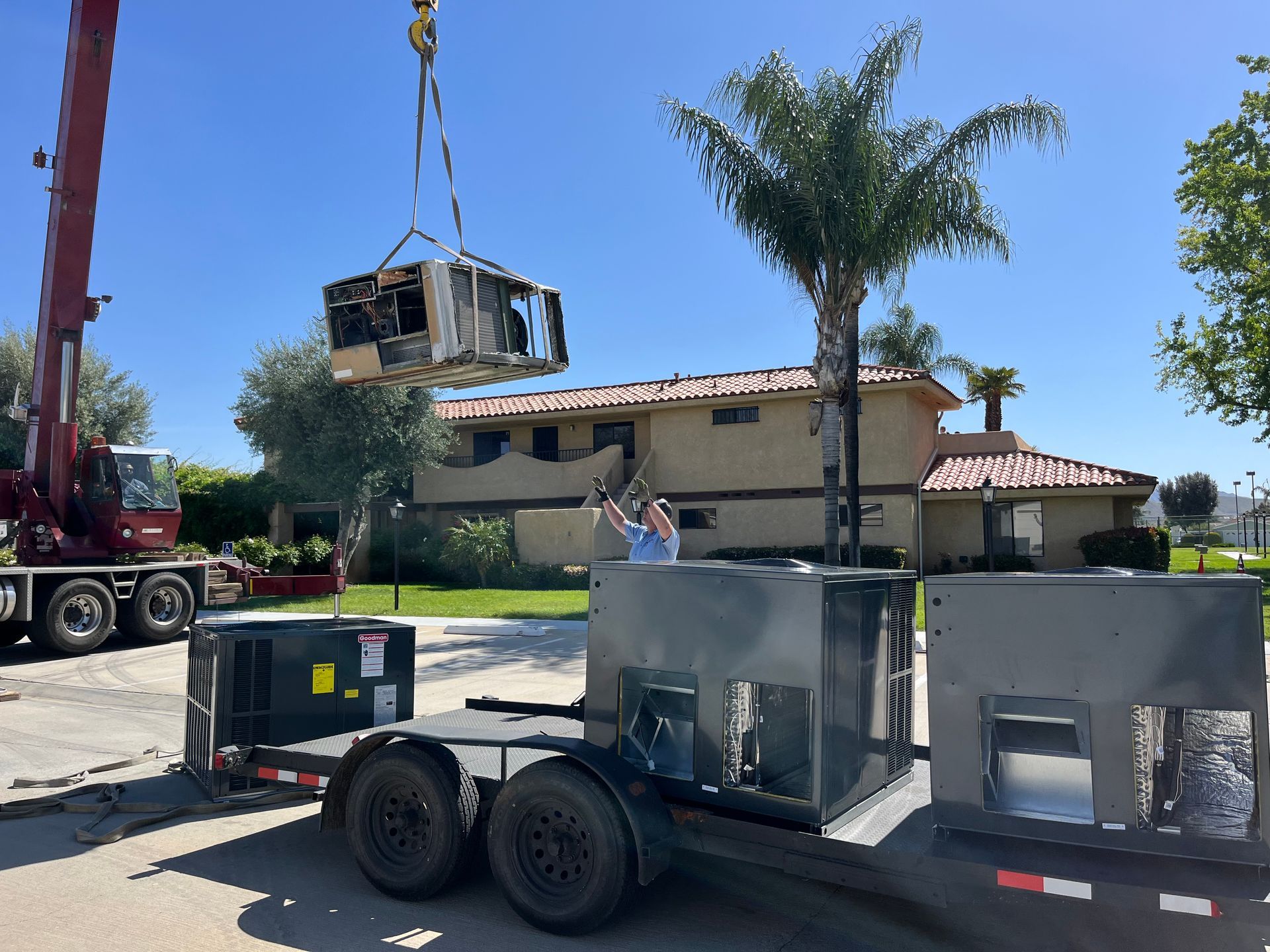 Crane lifting an AC unit over a trailer in front of a house; sunny day.