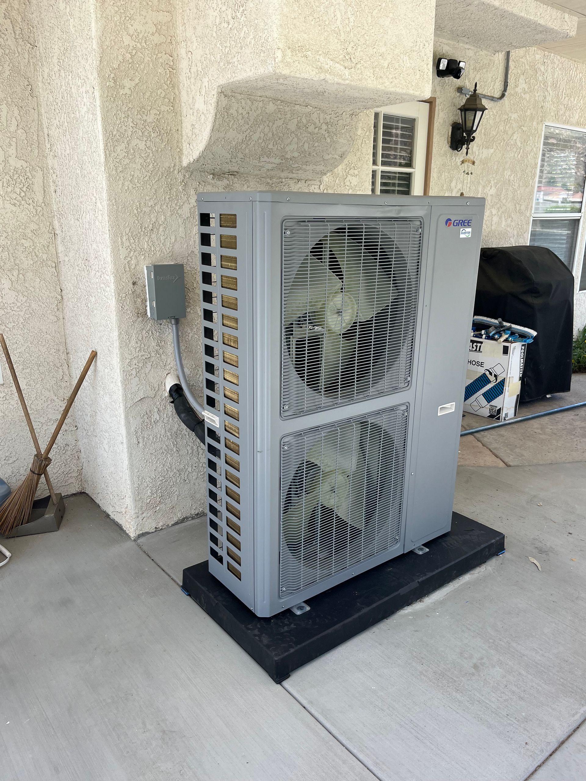 Gray air conditioning unit on a black pad, mounted on a concrete patio next to a stucco wall.