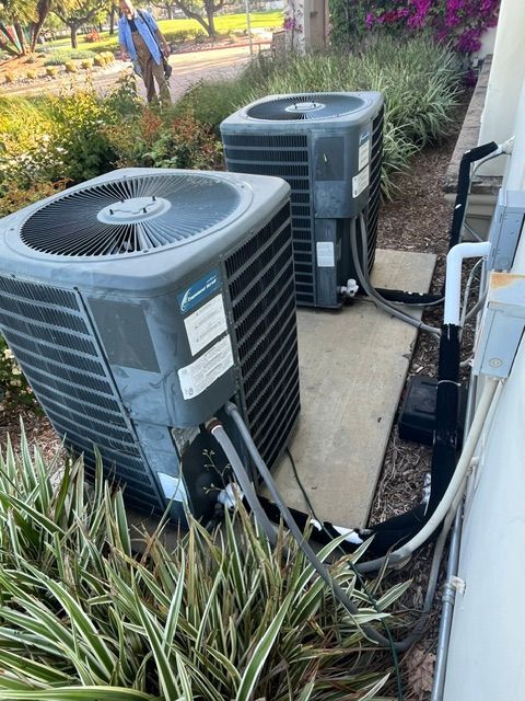 Two air conditioning units outside, surrounded by greenery and a person in the background.