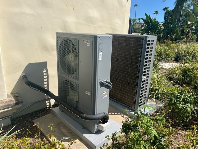 Two gray HVAC units outside a building, on concrete pads. Black tubes connect to the units, green plants surround them.