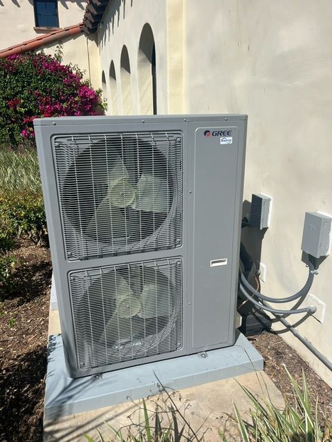 Exterior air conditioning unit, gray, mounted on a concrete pad beside a building, with two fan openings.