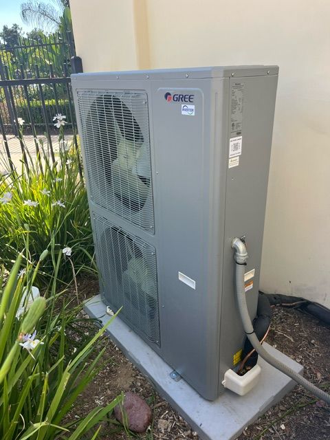 Gray heat pump unit outdoors, near plants, set on concrete.