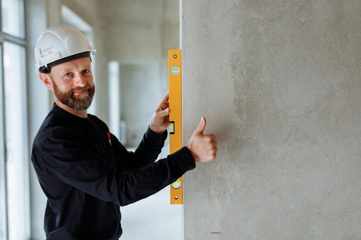 A worker wearing a hard hat holds a yellow spirit level against a concrete wall, giving a thumbs-up.