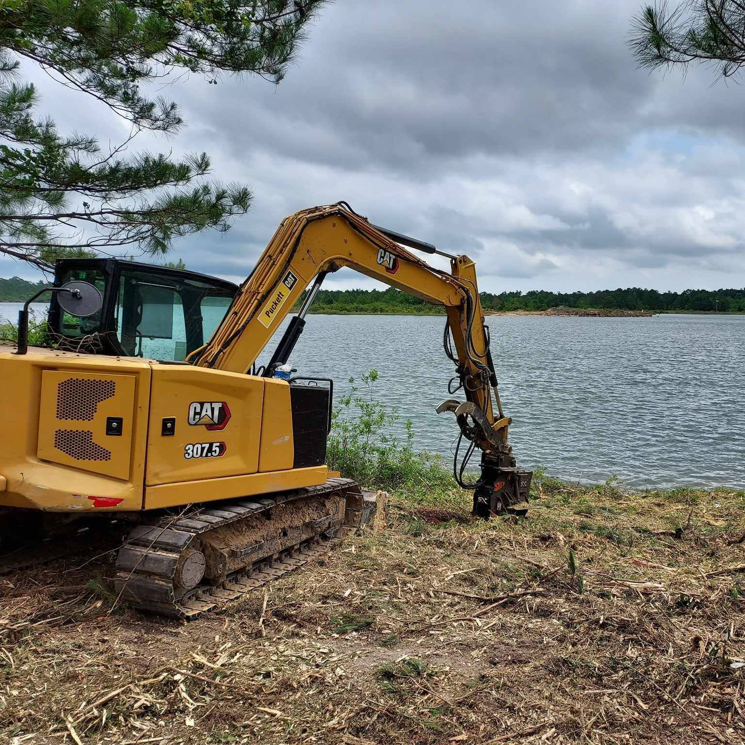 Yellow CAT excavator at lake edge, clearing vegetation under cloudy sky.