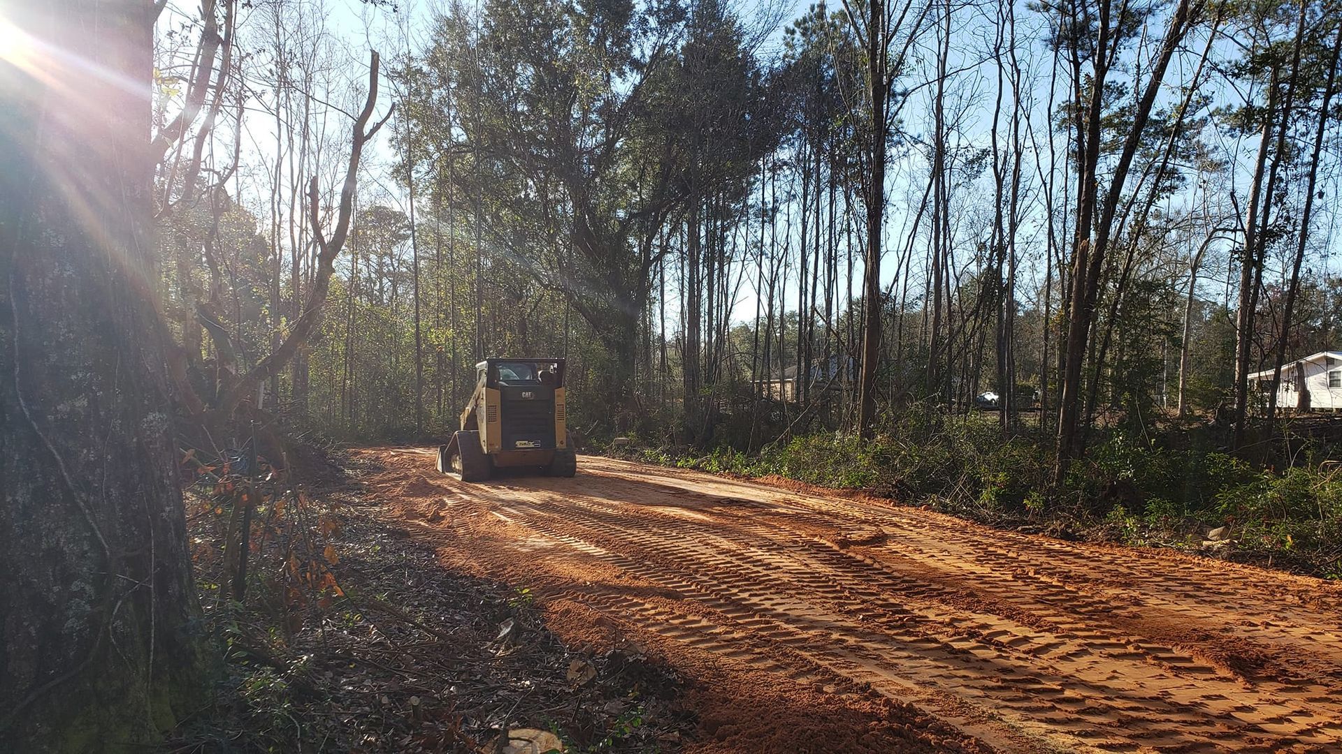 A skid steer clears a dirt road in a wooded area under a bright sky.