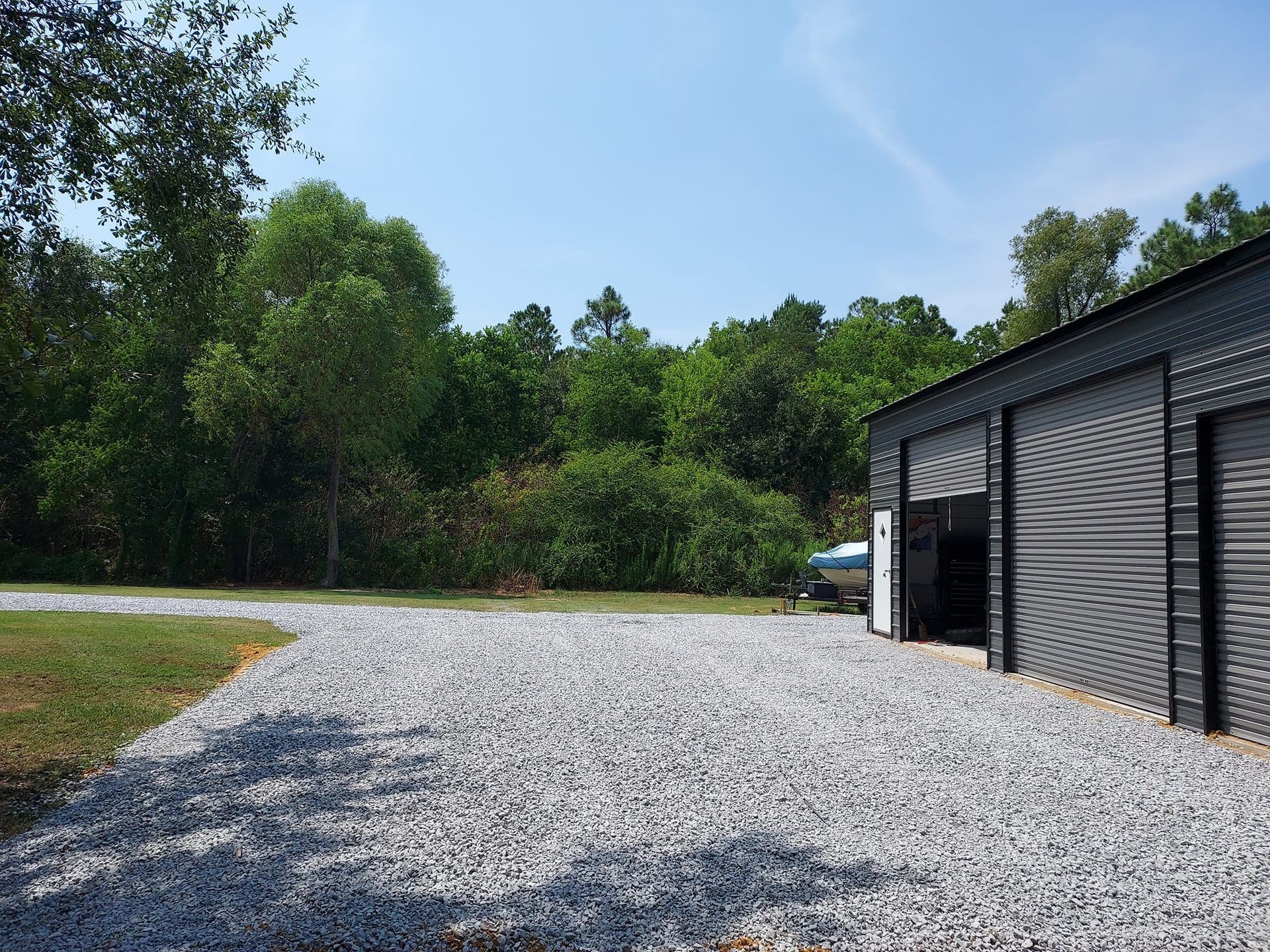 Gravel driveway leads to a dark grey garage with open doors. Trees and blue sky are in the background.