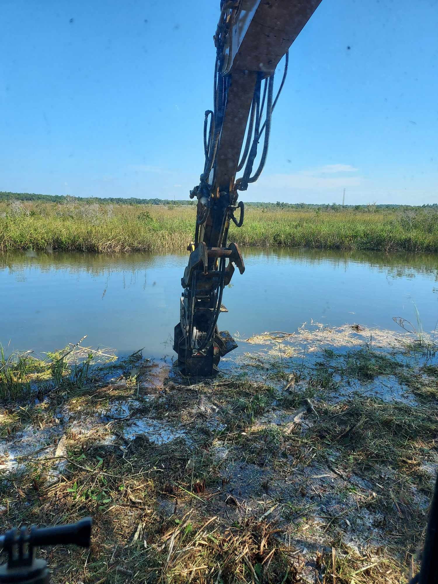 An excavator's arm digging into a body of water. The scene is outdoors with a blue sky.