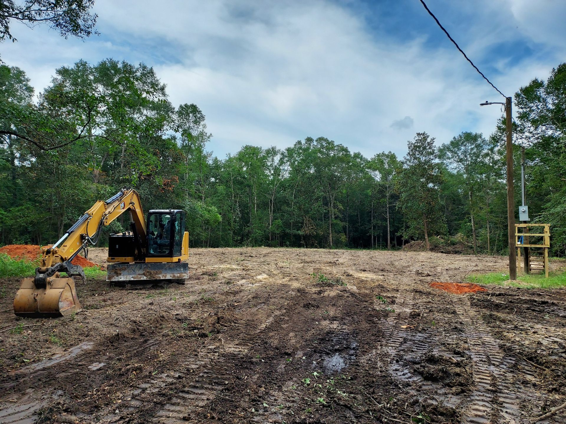 Excavator on a muddy, cleared lot. Trees in the background. Power pole on the right. Overcast sky.
