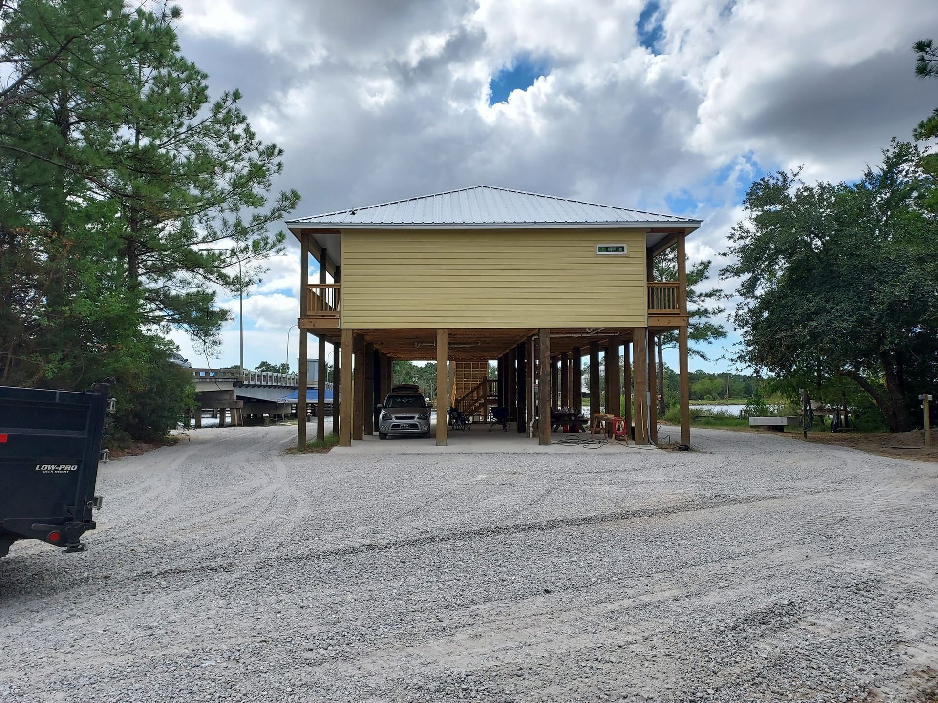 Yellow elevated house on stilts with car parked underneath. Gravel driveway and cloudy sky.