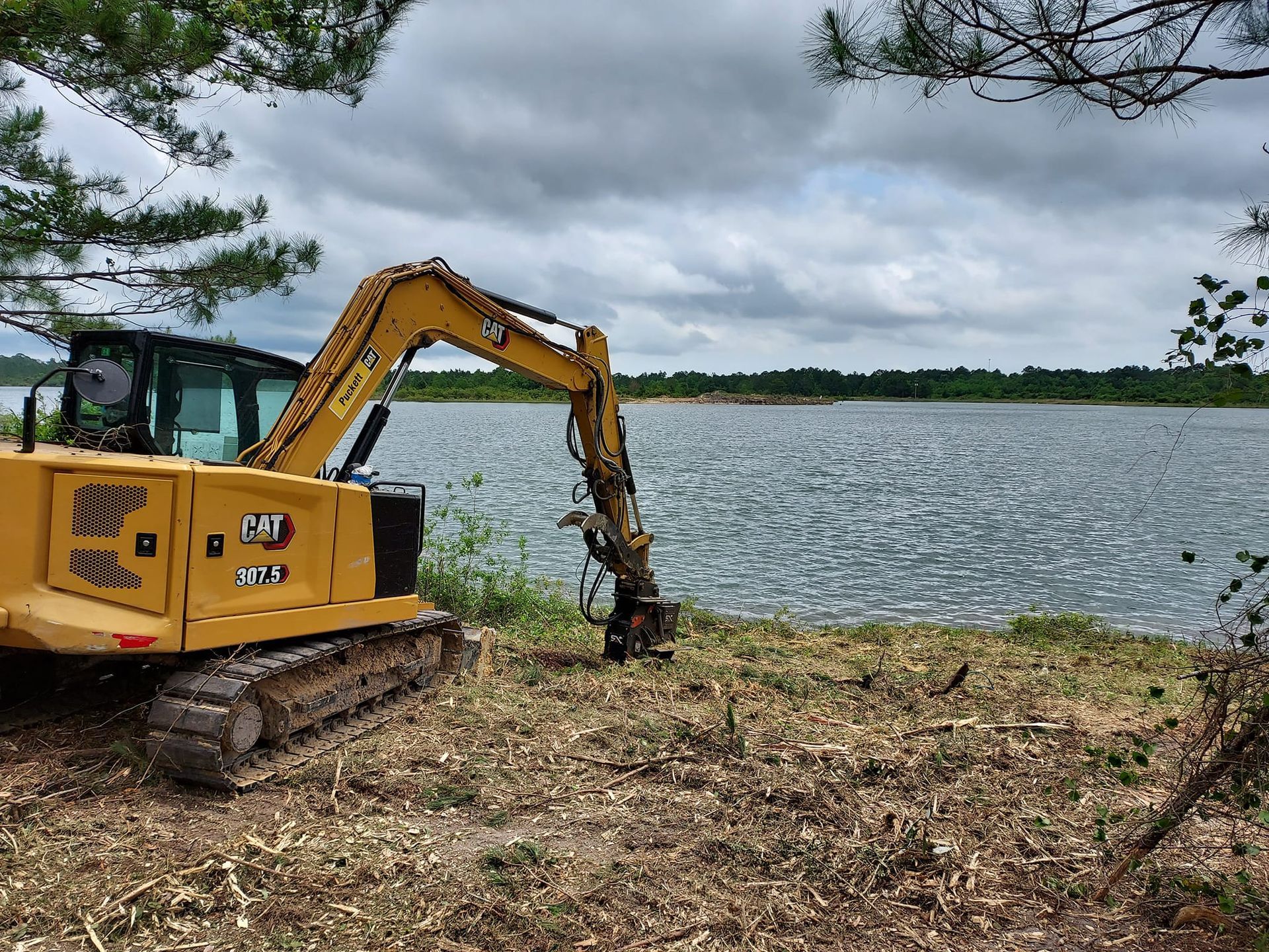 Yellow excavator near a lake, clearing brush under a cloudy sky.