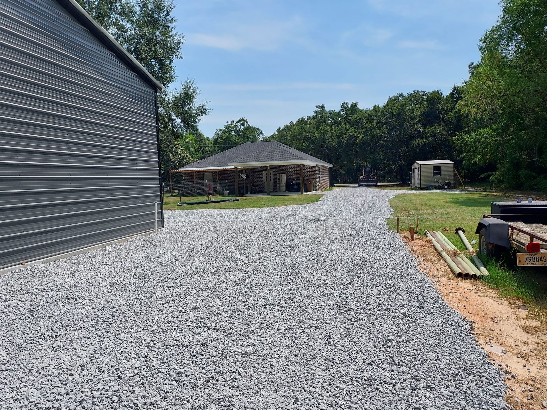 Gravel driveway leading to a brick house and shed. Gray metal building on the left, trees and blue sky in the background.