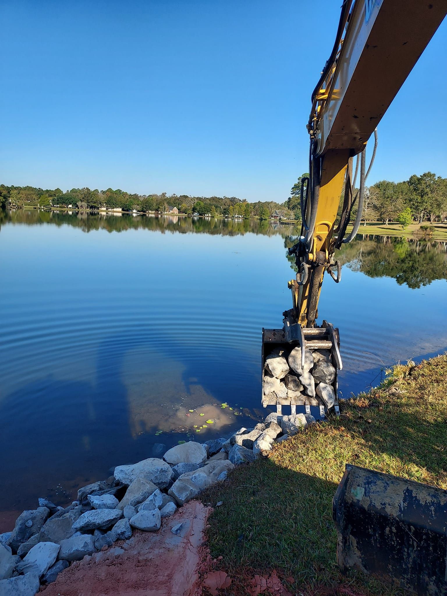 An excavator placing rocks along a lake's edge under a clear, blue sky.