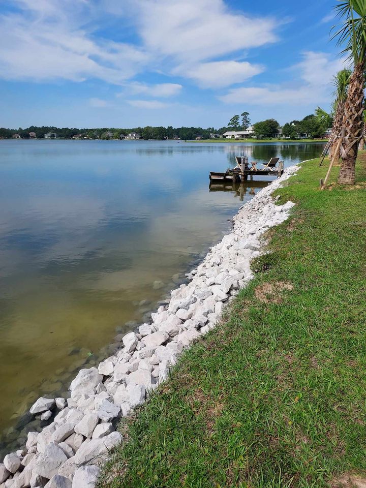 Lakeside view: rocky shoreline, calm water reflecting a blue sky, small dock, green grass, and trees.