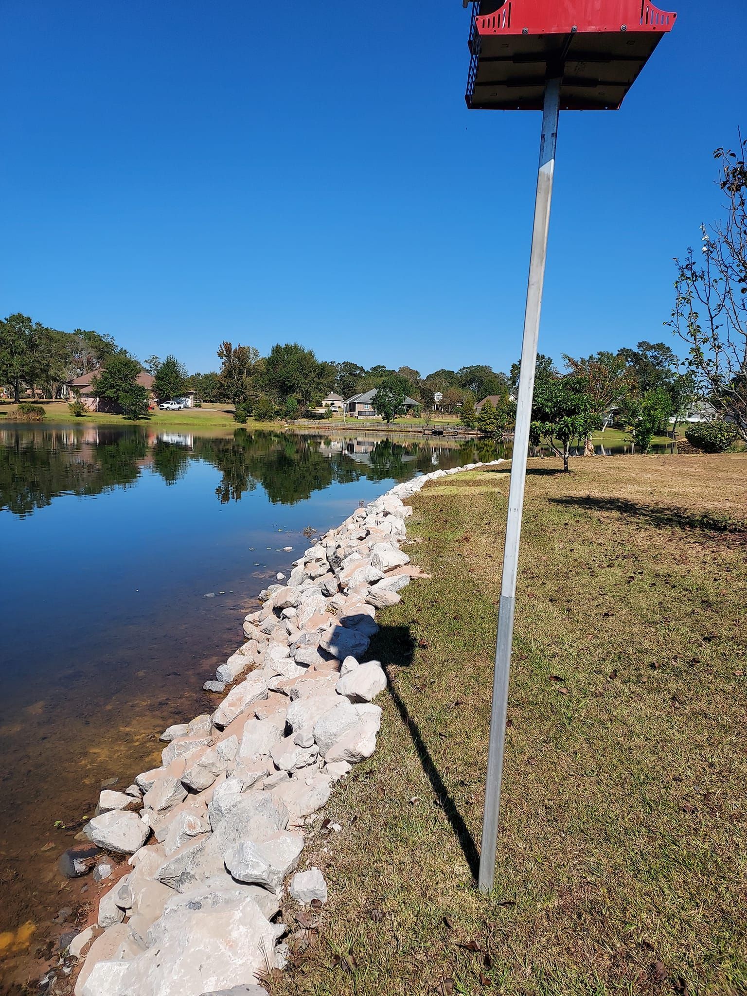 Birdhouse on a pole by a lake with a rocky shoreline and a row of houses in the distance on a sunny day.