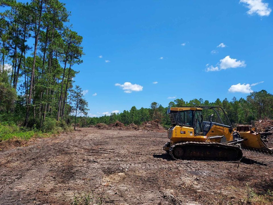 A bulldozer on a cleared dirt lot next to a forest, under a blue sky with white clouds.