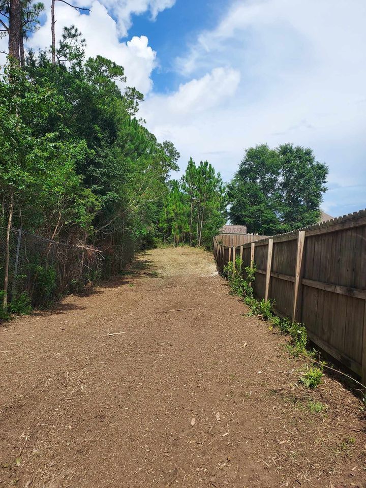 A cleared path between a fence and trees under a partly cloudy sky.