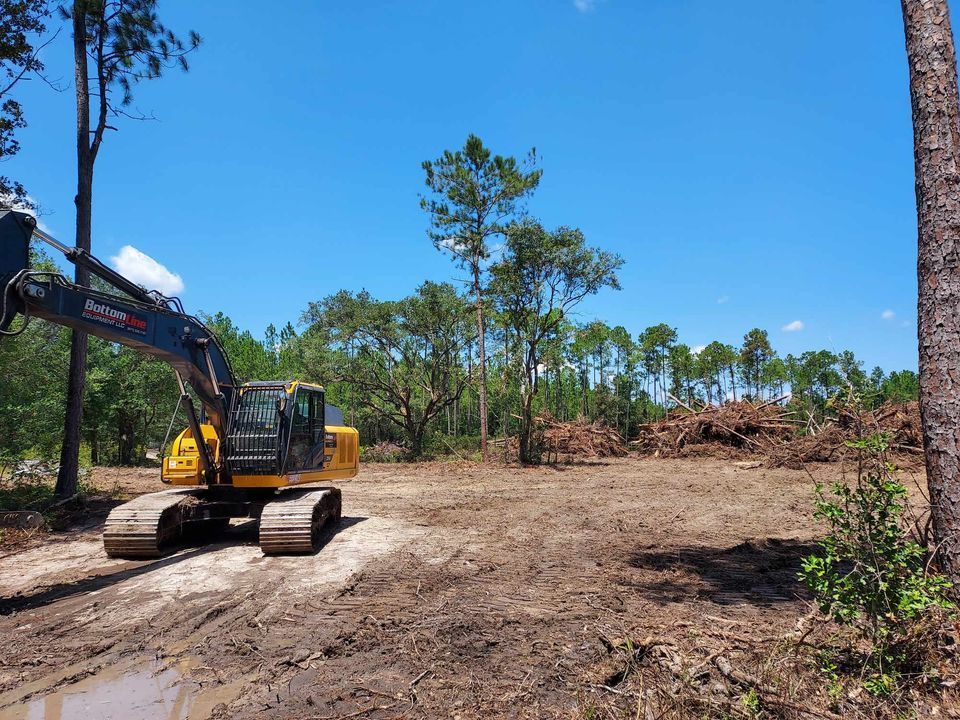 Yellow excavator on muddy ground clearing a forest under a blue sky.