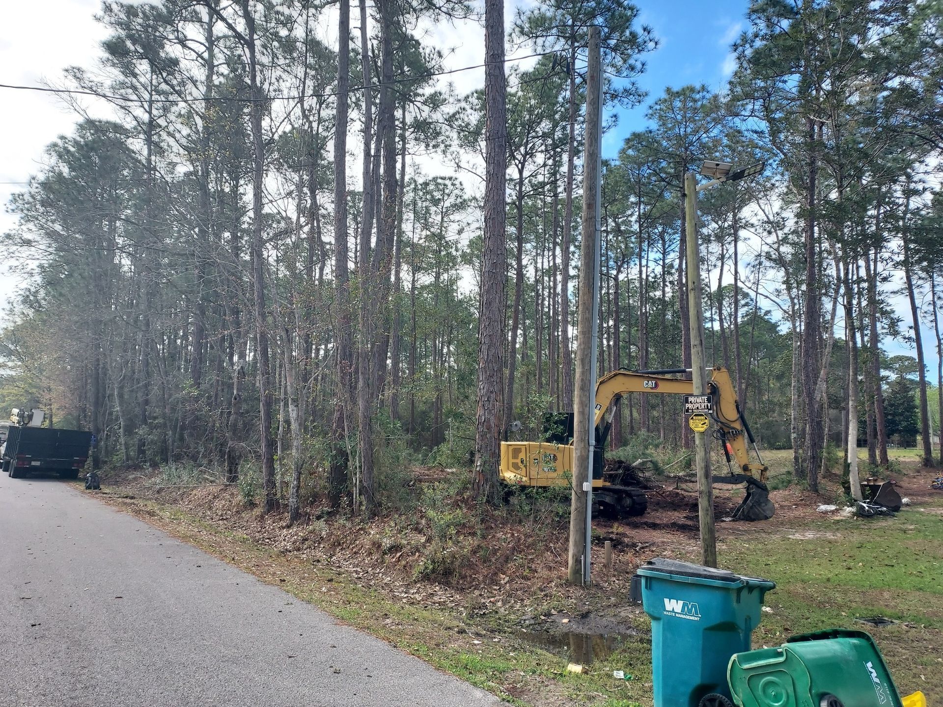 Yellow excavator clearing trees near a street with trash cans, a utility pole, and a gray road.