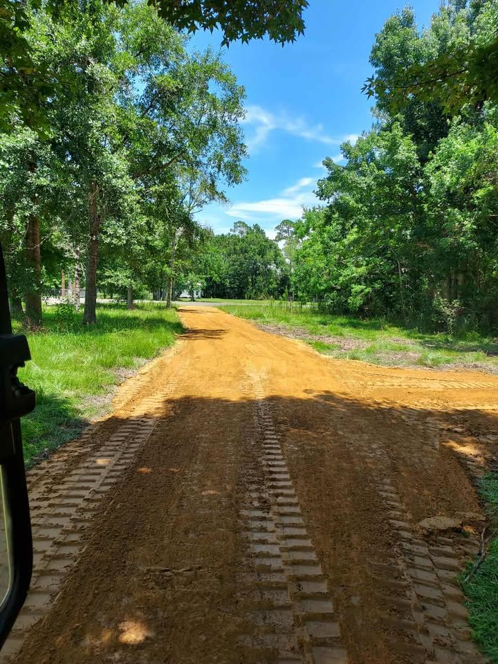 Dirt road lined with green trees under a blue sky, showing construction work in progress.