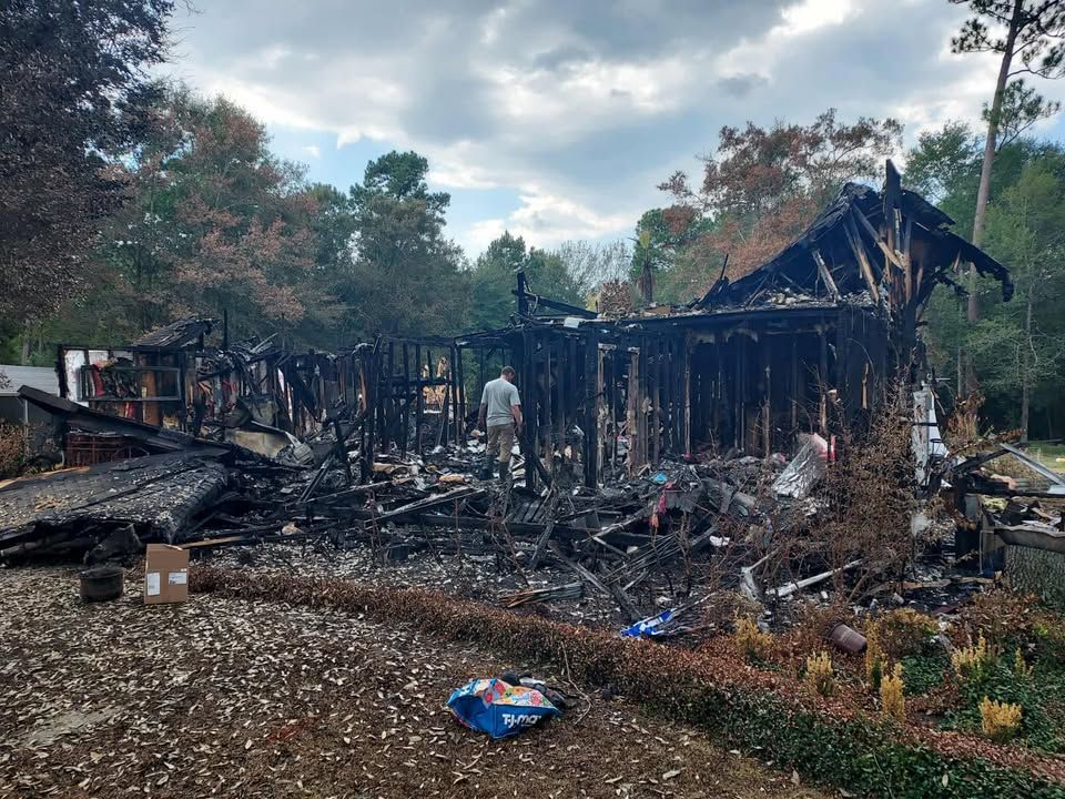 A charred, burnt-out house with a person surveying the damage in a wooded area under a cloudy sky.