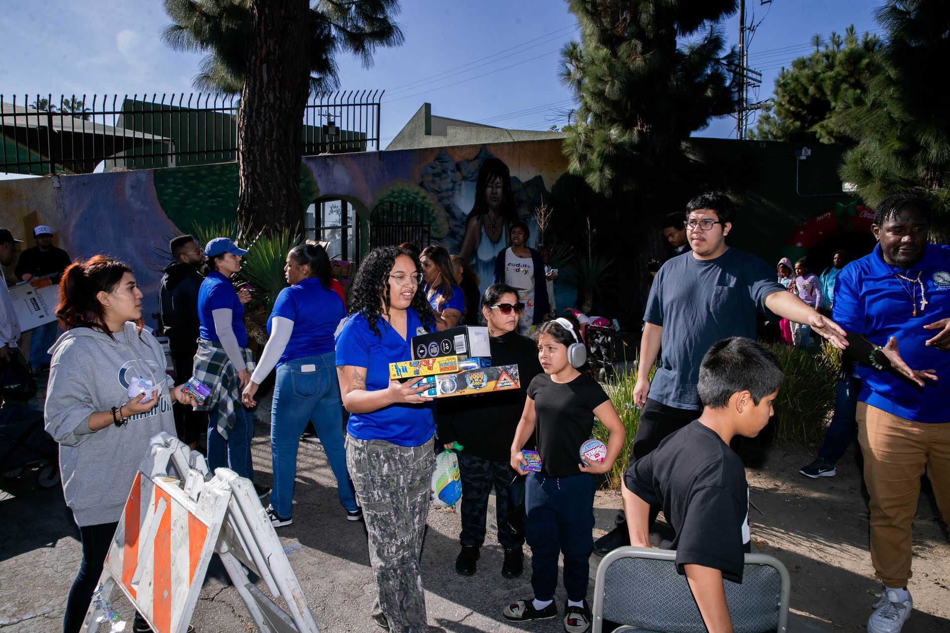 People distributing items outdoors; some wear blue shirts.  Children and adults gathered near a wall.