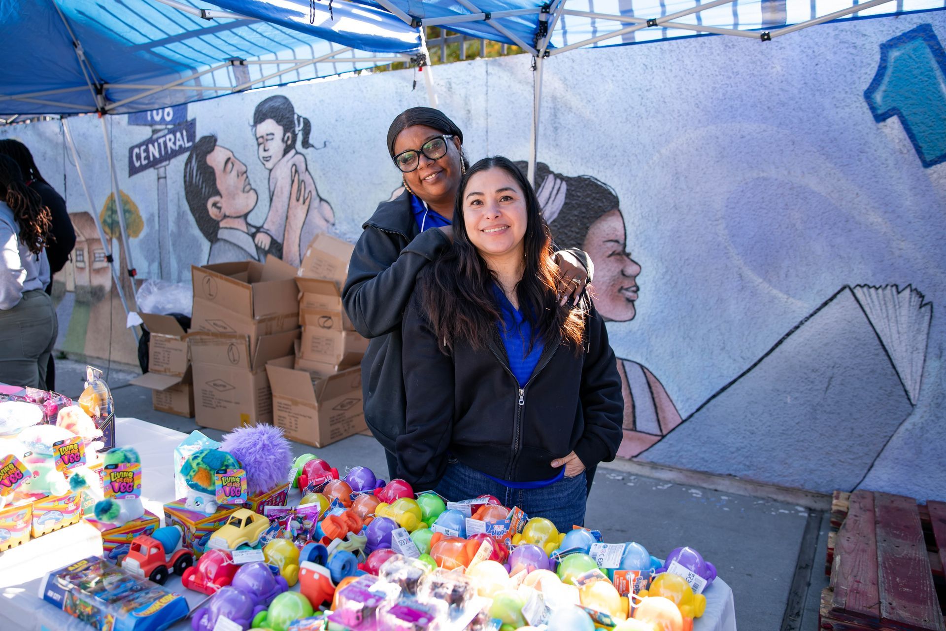 Two people smiling at a table with toys under a blue canopy; mural in the background.