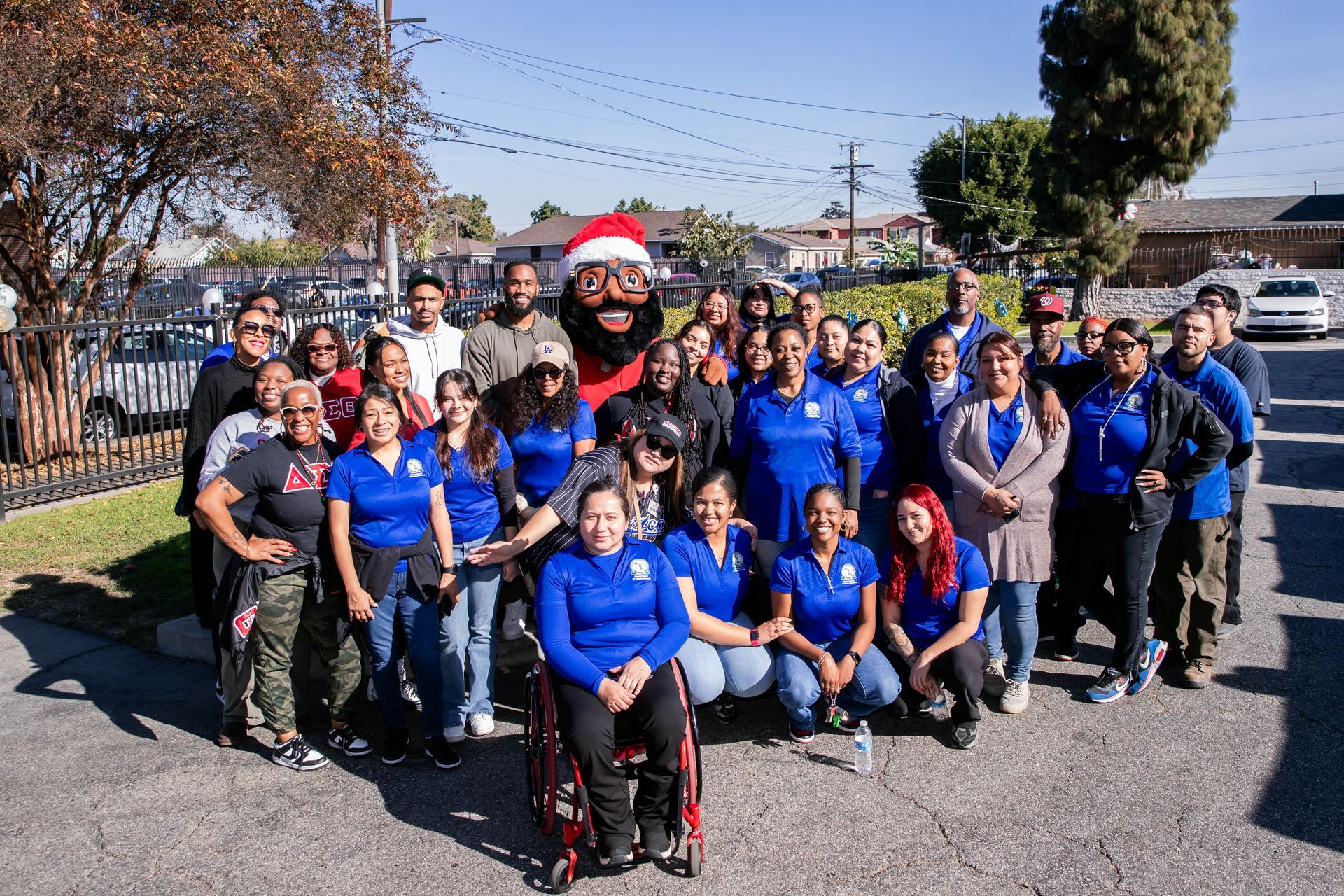 Group of people with a large Santa head in a sunny outdoor setting, some wearing blue shirts.
