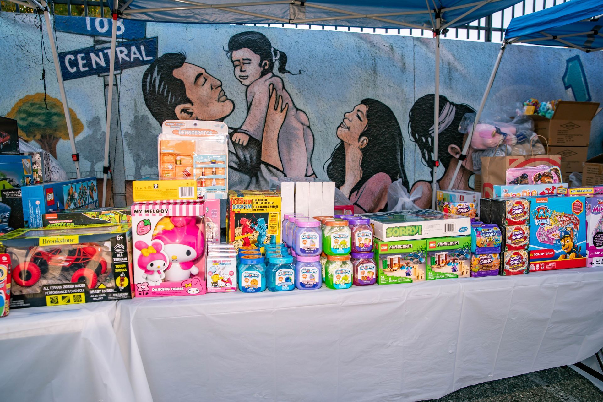 Toys displayed on a table at an outdoor market, with a mural of people in the background.