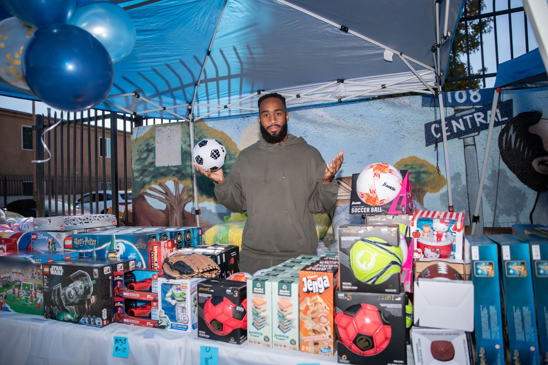 John Johnson III at table with toys and soccer balls. Blue and white balloons, outdoor setting.