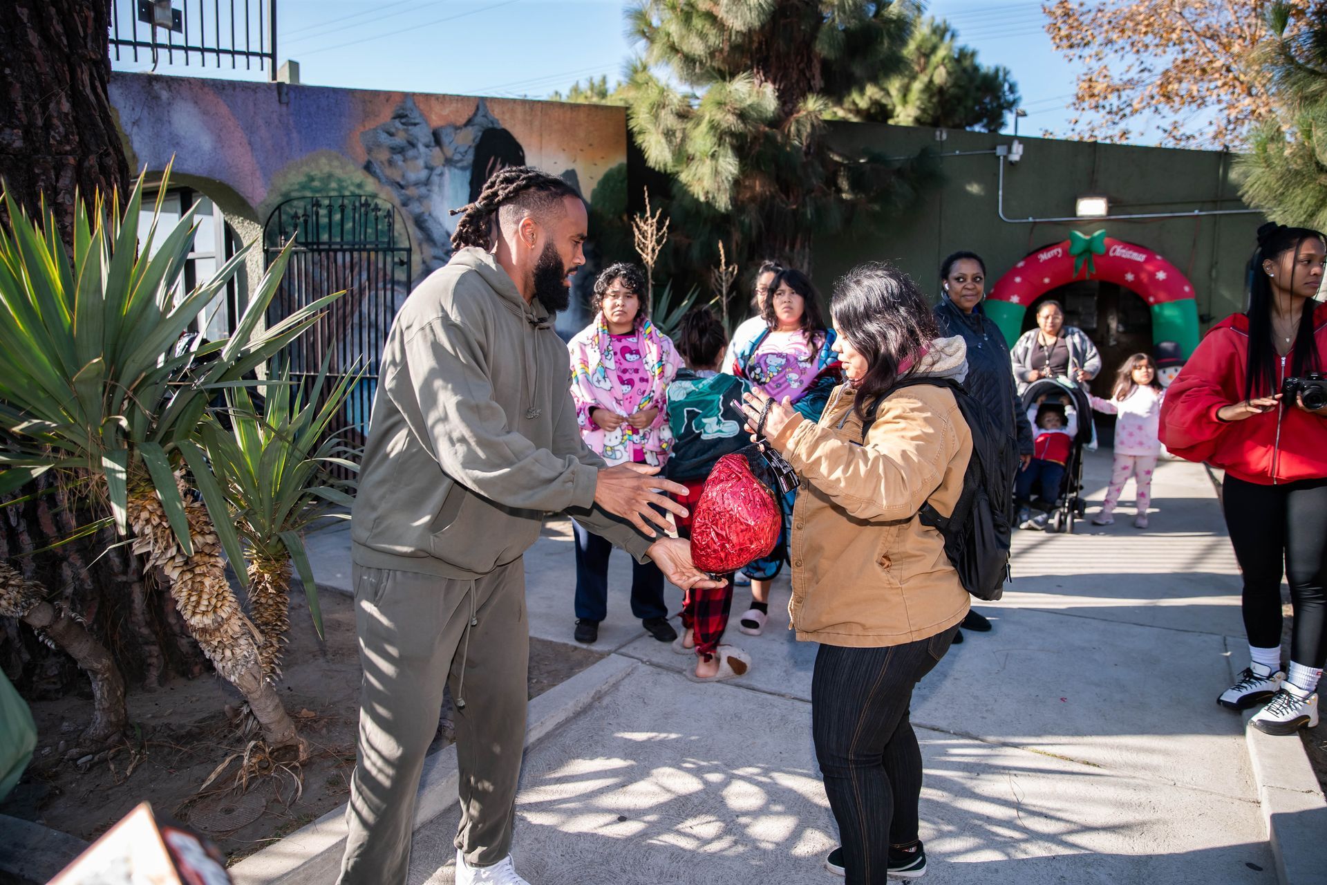 John Johnson III in gray sweatsuit handing a gift to a woman outside. Children and other adults are present.