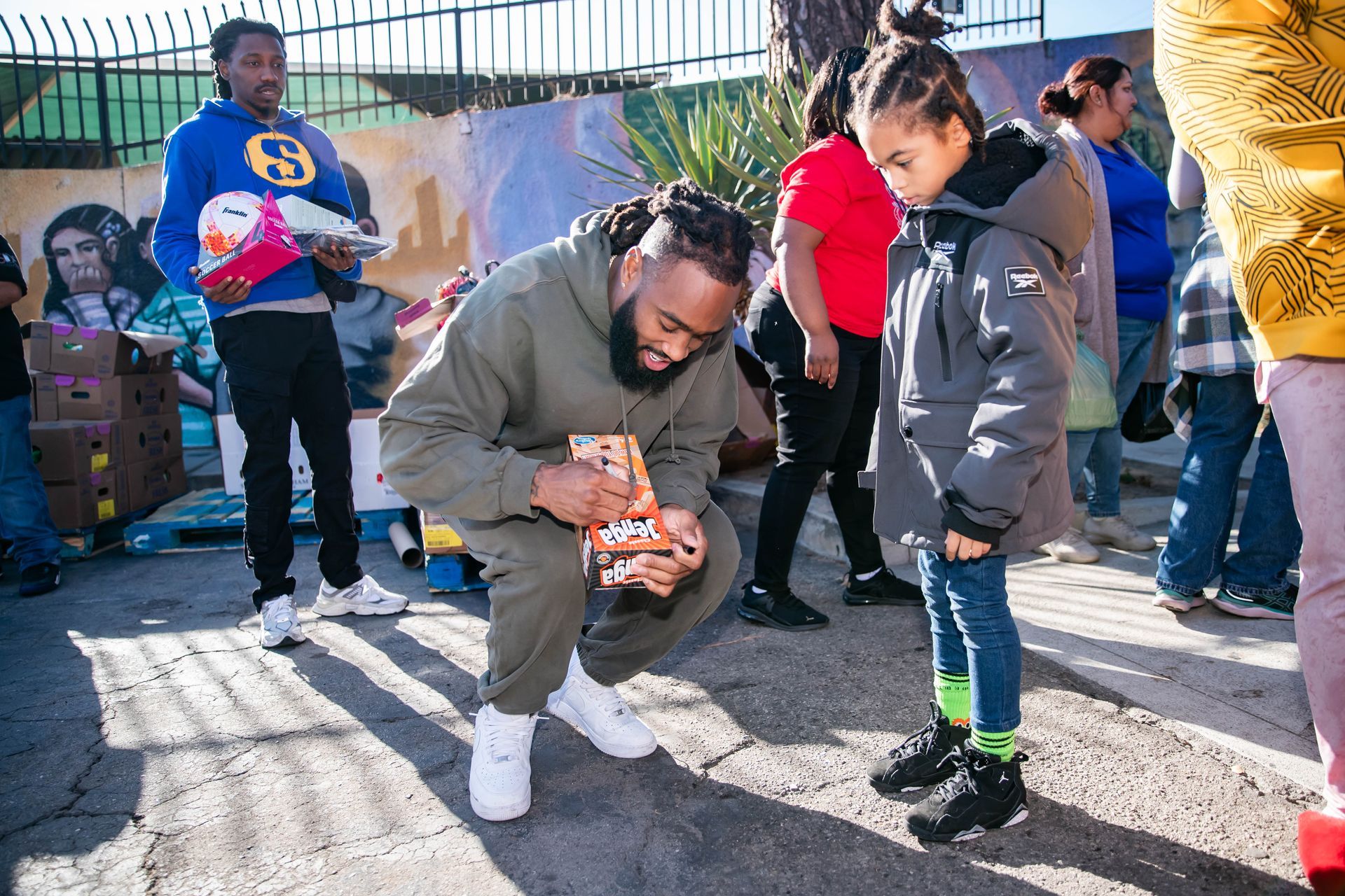 John Johnson III kneeling, opening gift for a child outdoors near crowd and mural.