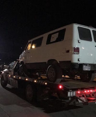 A white van is being towed by a tow truck at night.