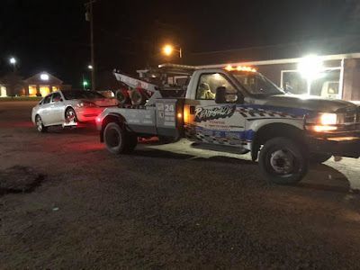 A tow truck is towing a car in a parking lot at night.