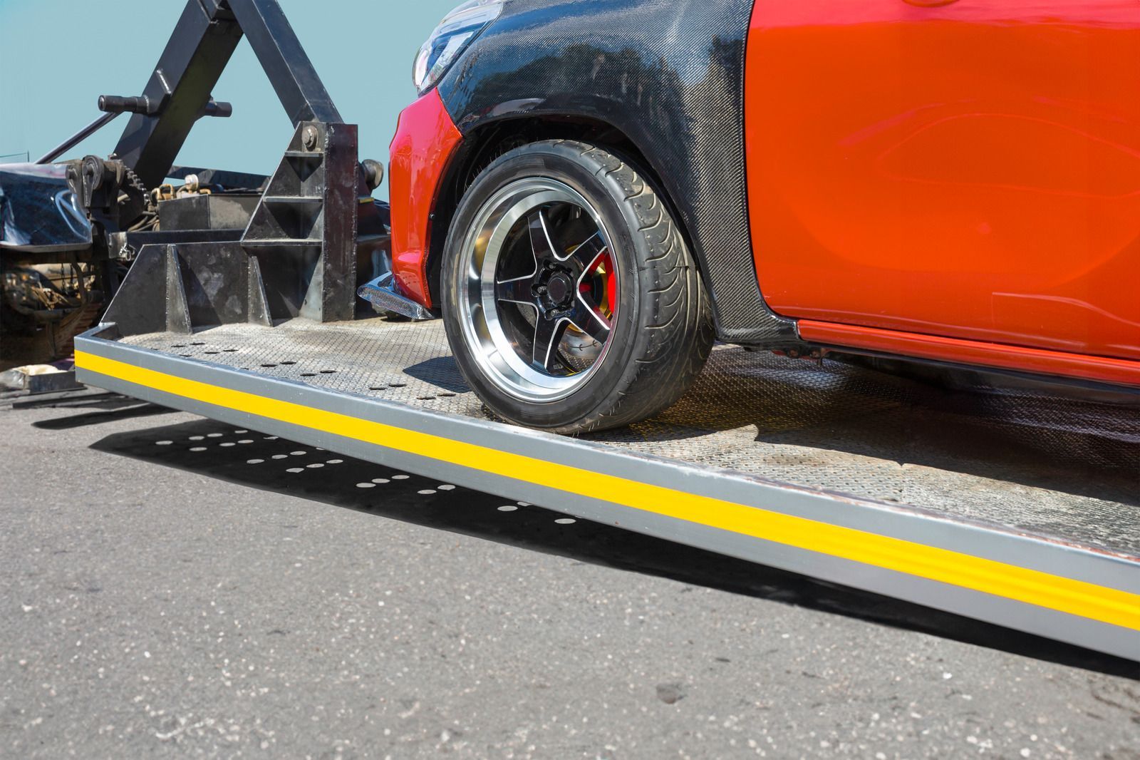 Red car being towed on a flatbed truck; close-up on wheel and ramp, outdoors.