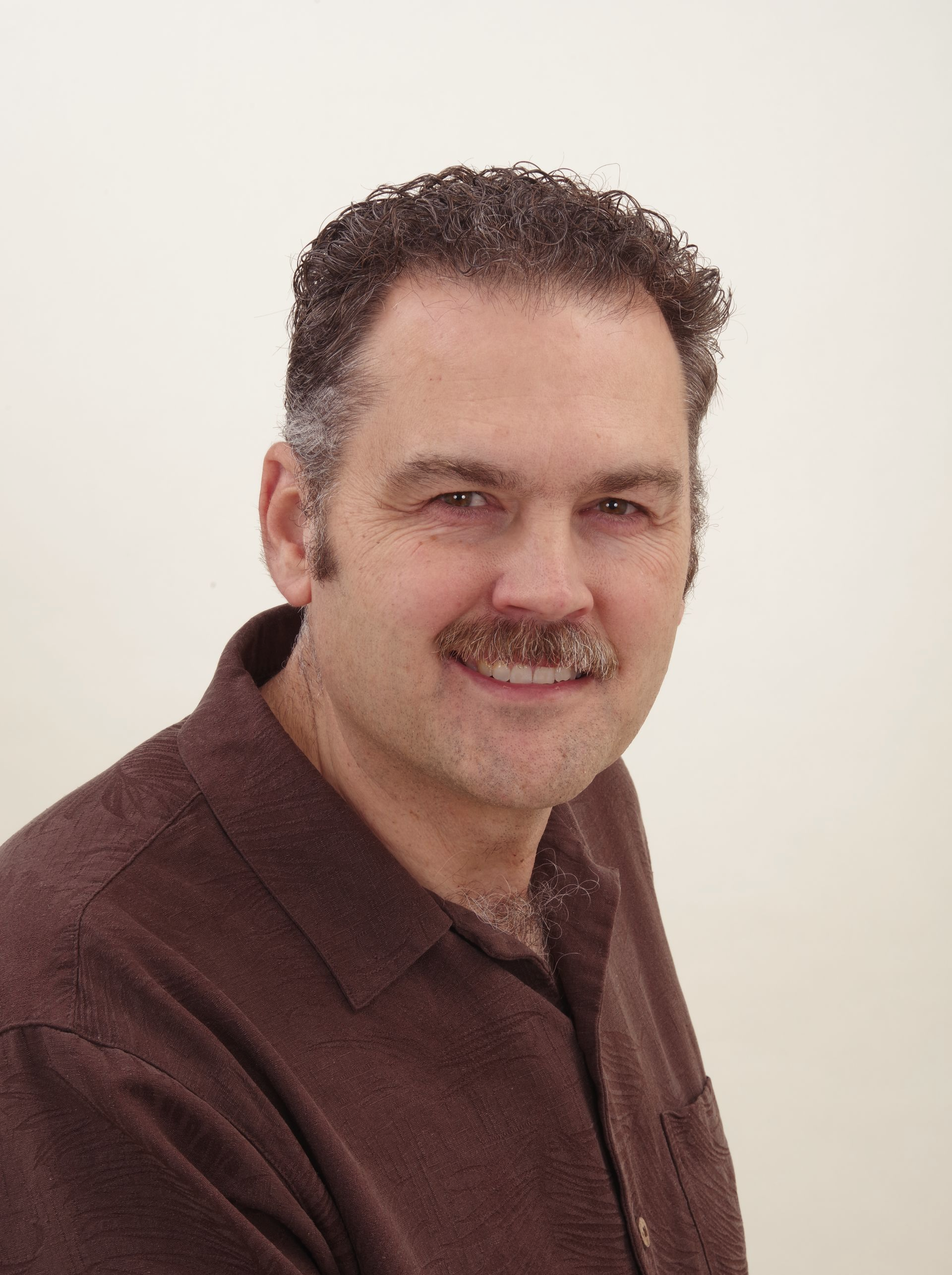 A man with a mustache is wearing a brown shirt and smiling.