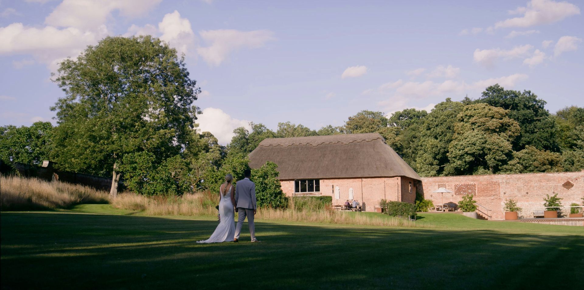 A bride and groom are walking in a field in front of a thatched barn.