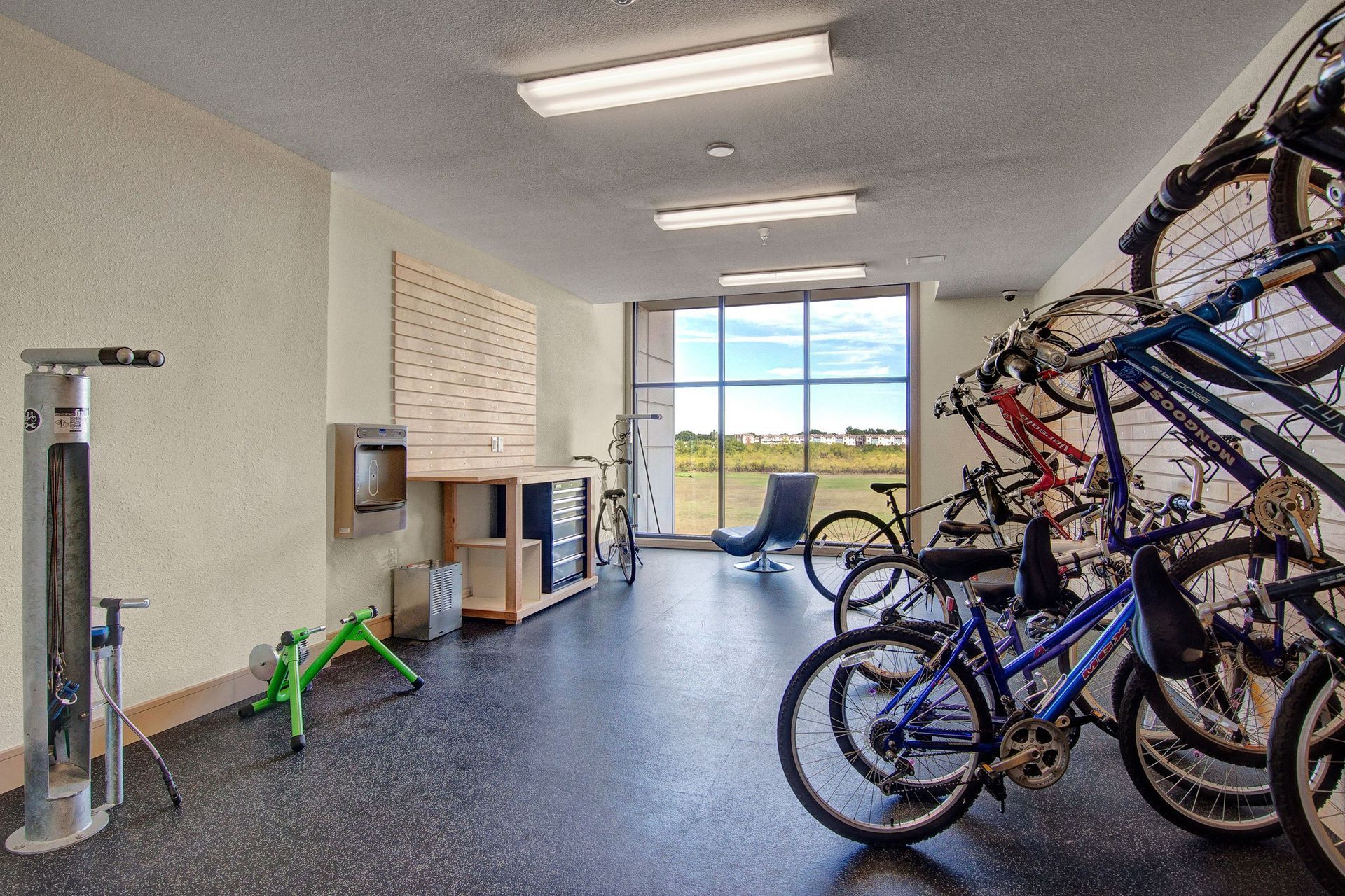 A bright bike repair room here at The Residence at The COR Apartment Homes with bikes on vertical racks, tools on a workbench, and a green bike stand. Large windows offer a scenic outdoor view.