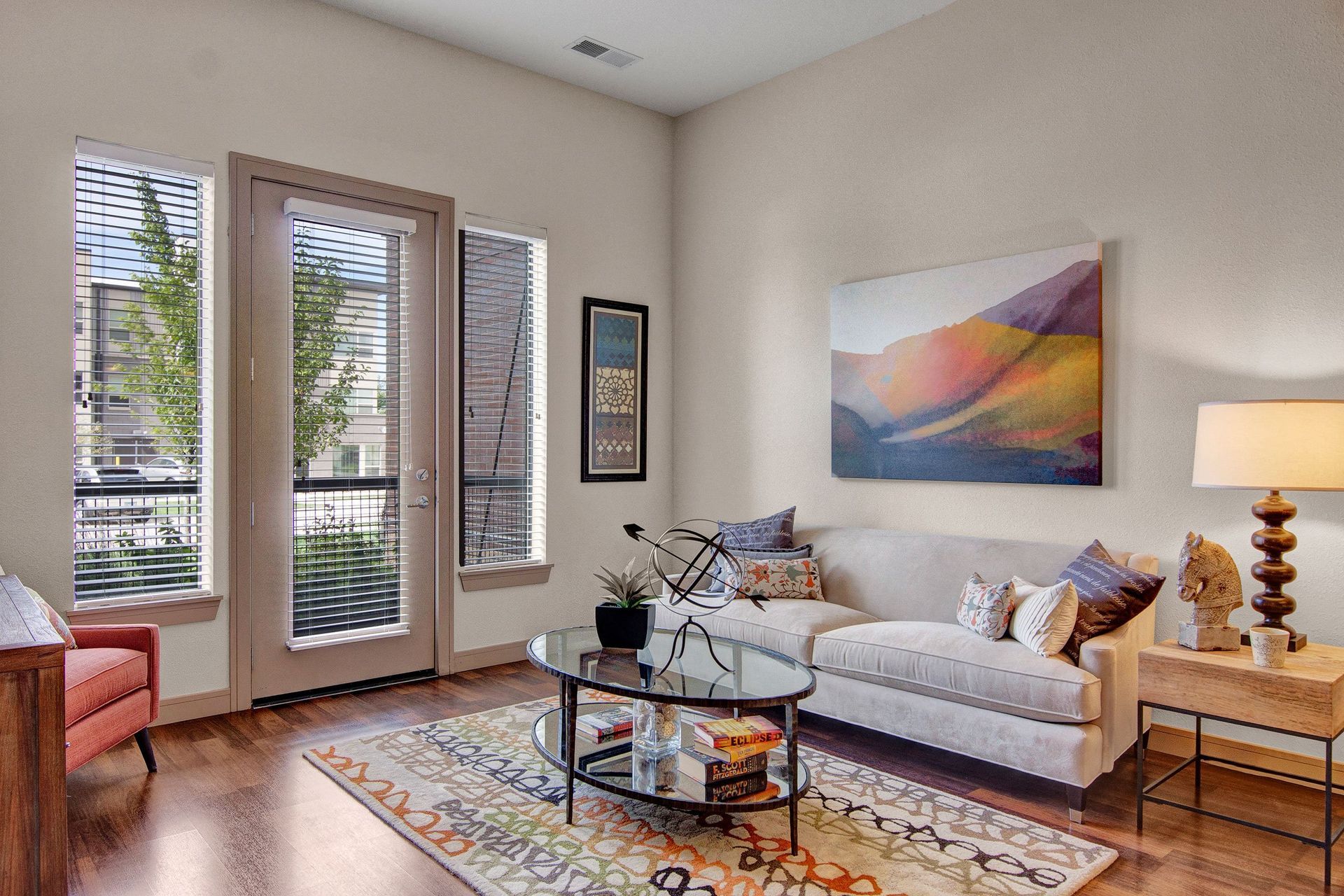 Modern living room here at The Residence at The COR Apartment Homes with a beige sofa, colorful cushions, and a landscape painting. A glass coffee table holds books, and a lamp lights a wooden side table.