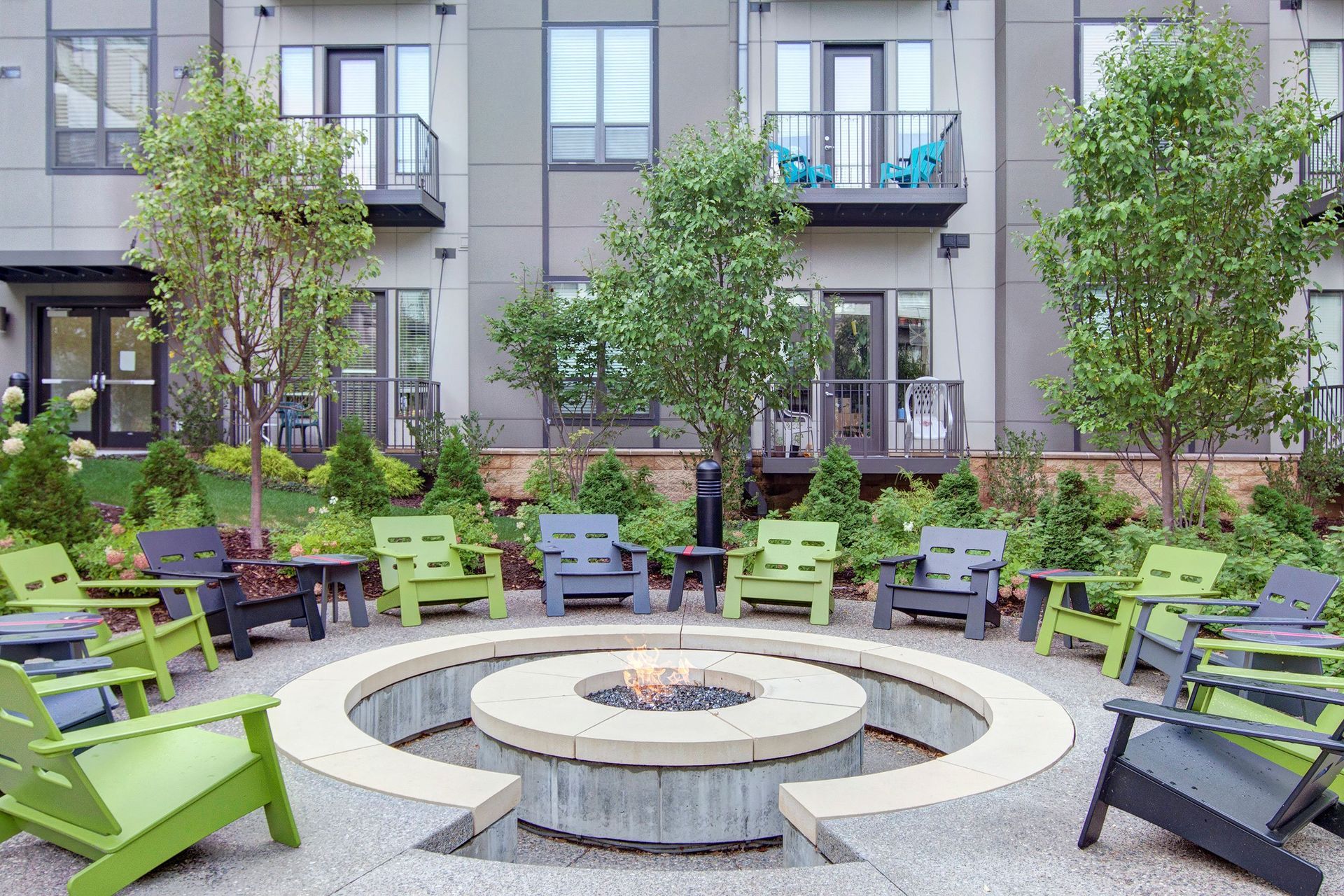 Circular fire pit here at The Residence at The COR Apartment Homes with a gentle flame, surrounded by green and black chairs on a patio. Modern buildings and lush greenery create a serene atmosphere.