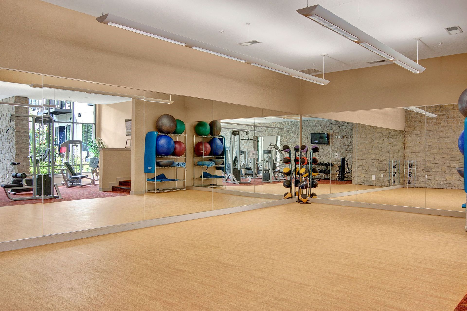 Spacious gym here at The Residence at The COR Apartment Homes with mirrored walls, exercise balls on shelves, and a dumbbell rack. Natural light from large windows creates an inviting atmosphere.