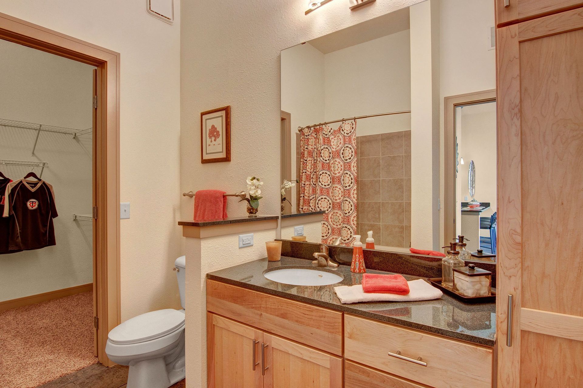 Cozy bathroom here at The Residence at The COR Apartment Homes with wood cabinetry, a black granite countertop with a sink, red accents, a decorative shower curtain, and view into a closet with jerseys.