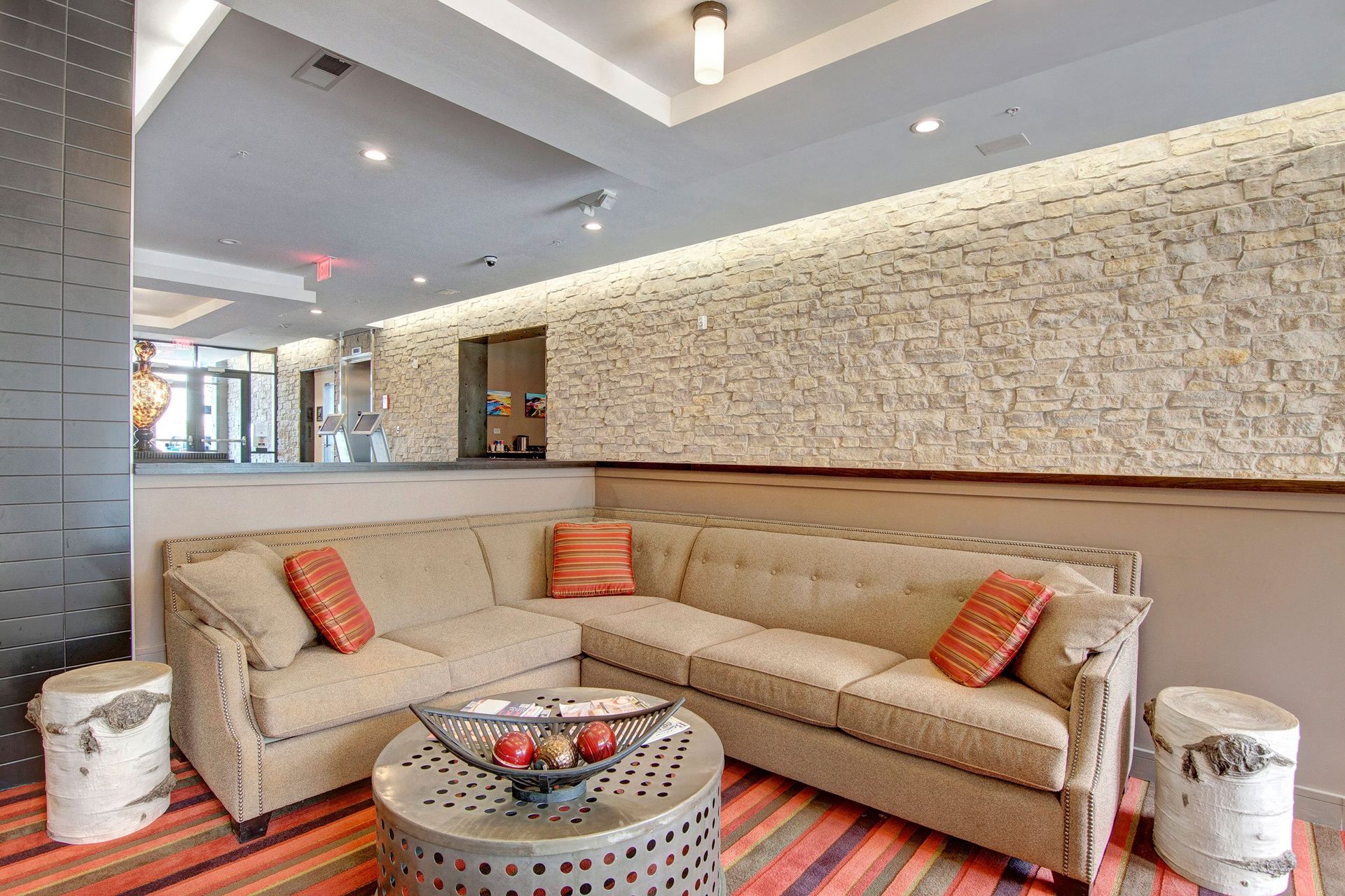 Modern lounge here at The Residence at The COR Apartment Homes with a beige sectional sofa, colorful striped pillows, and a metal coffee table topped with decorative spheres. Stone wall and warm lighting create a cozy atmosphere.