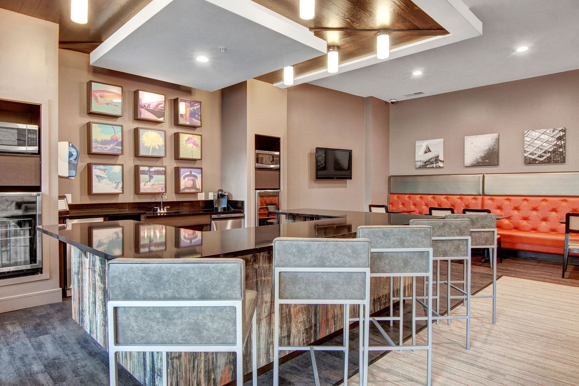 Contemporary kitchen space here at The Residence at The COR Apartment Homes with a rustic island, tall chairs, framed artwork on walls, modern lighting, and orange tufted seating. Warm and inviting tone.