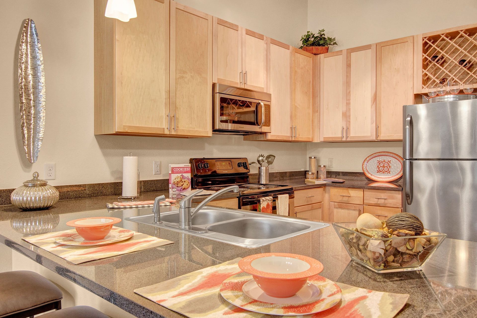 Modern kitchen here at The Residence at The COR Apartment Homes with wooden cabinets, stainless steel appliances, and a glossy countertop. Decor includes a fruit bowl, colorful dishes, and a cookbook, creating a warm, inviting atmosphere.