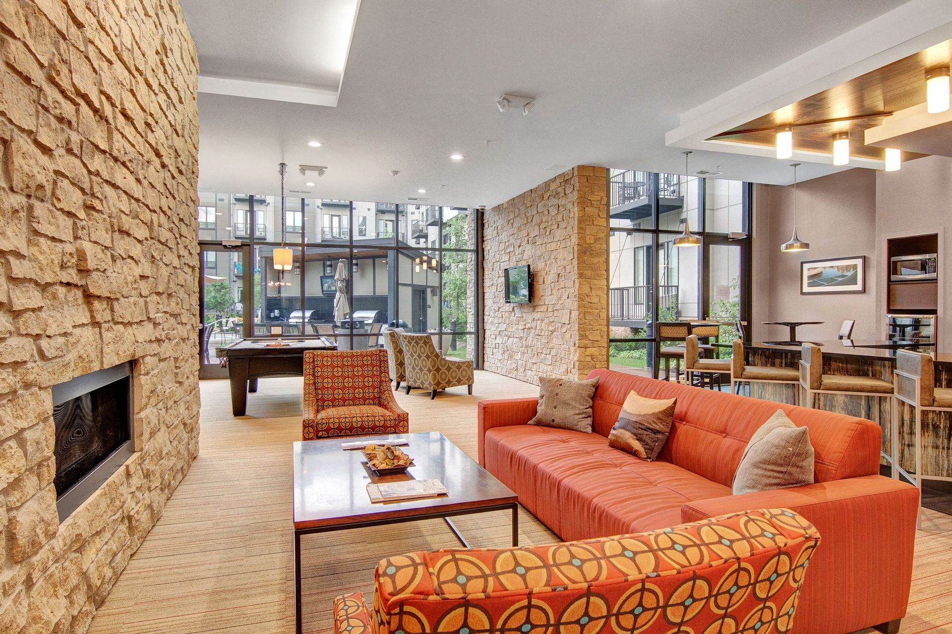 Modern lobby here at The Residence at The COR Apartment Homes with stone walls featuring a cozy red sofa, patterned armchairs, and a pool table. Large windows offer a view of the courtyard. Warm, inviting ambiance.