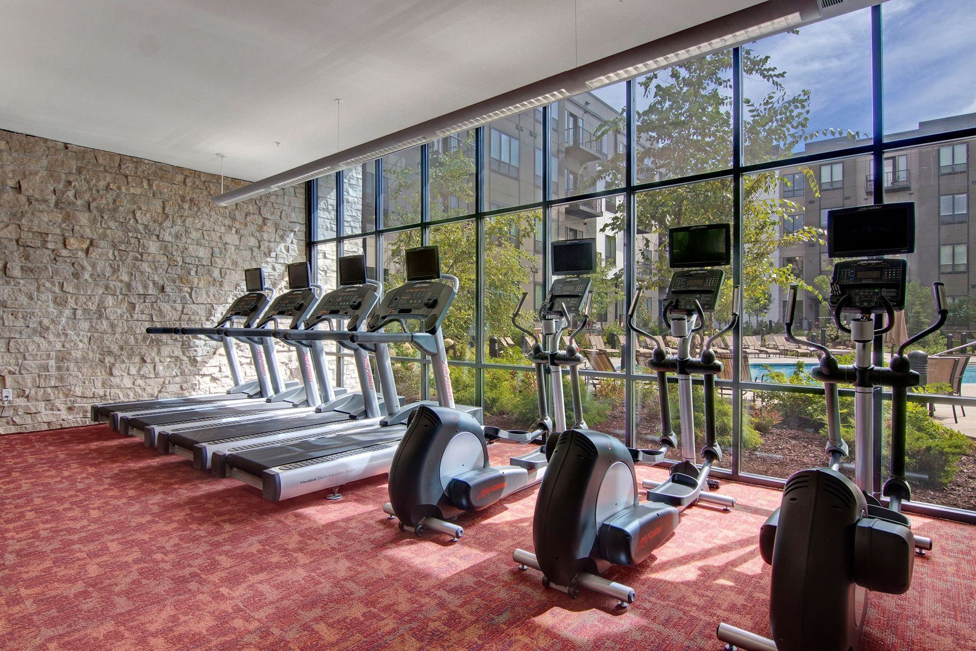 Sunlit gym here at The Residence at The COR Apartment Homes with treadmills and ellipticals lined up against large windows. Outside, trees and buildings are visible, creating an inviting atmosphere.