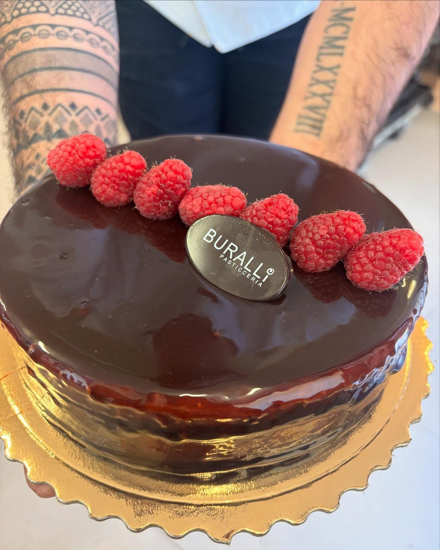 Un uomo con un tatuaggio sul braccio tiene in mano una torta al cioccolato con lamponi sopra