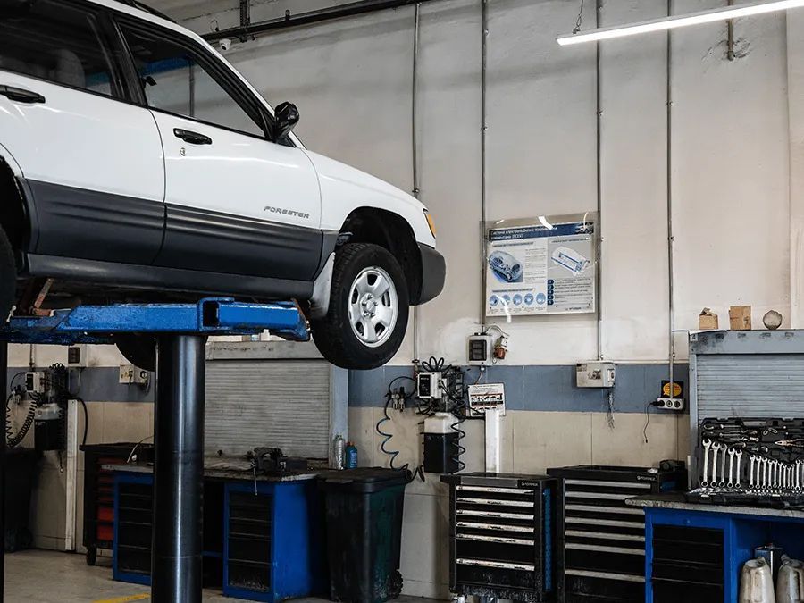 White car on a hydraulic lift in a repair shop. Tools and equipment visible.