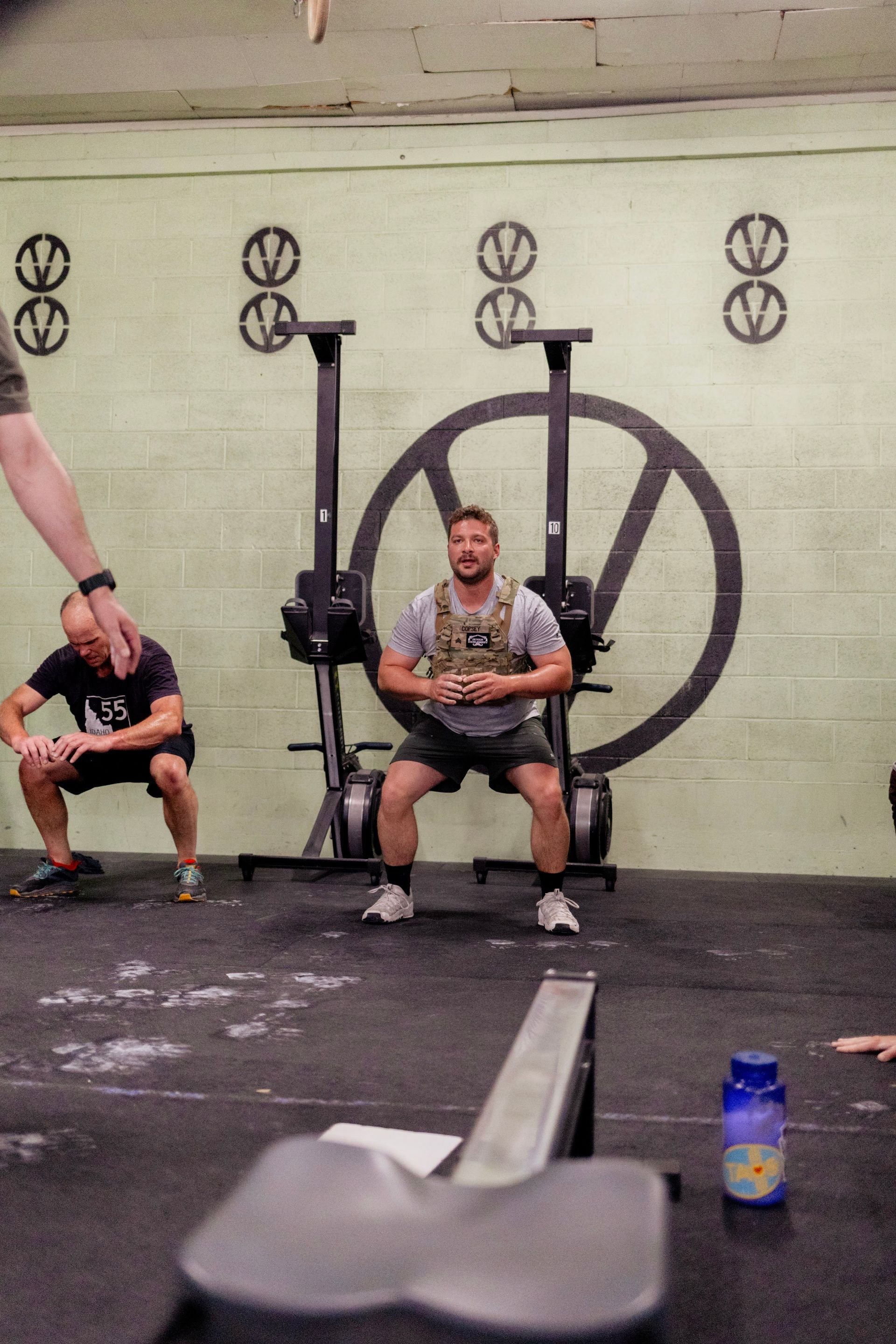 Man squats at Verdant CrossFit gym in Boise wearing a weighted vest. Another man in the background assists.