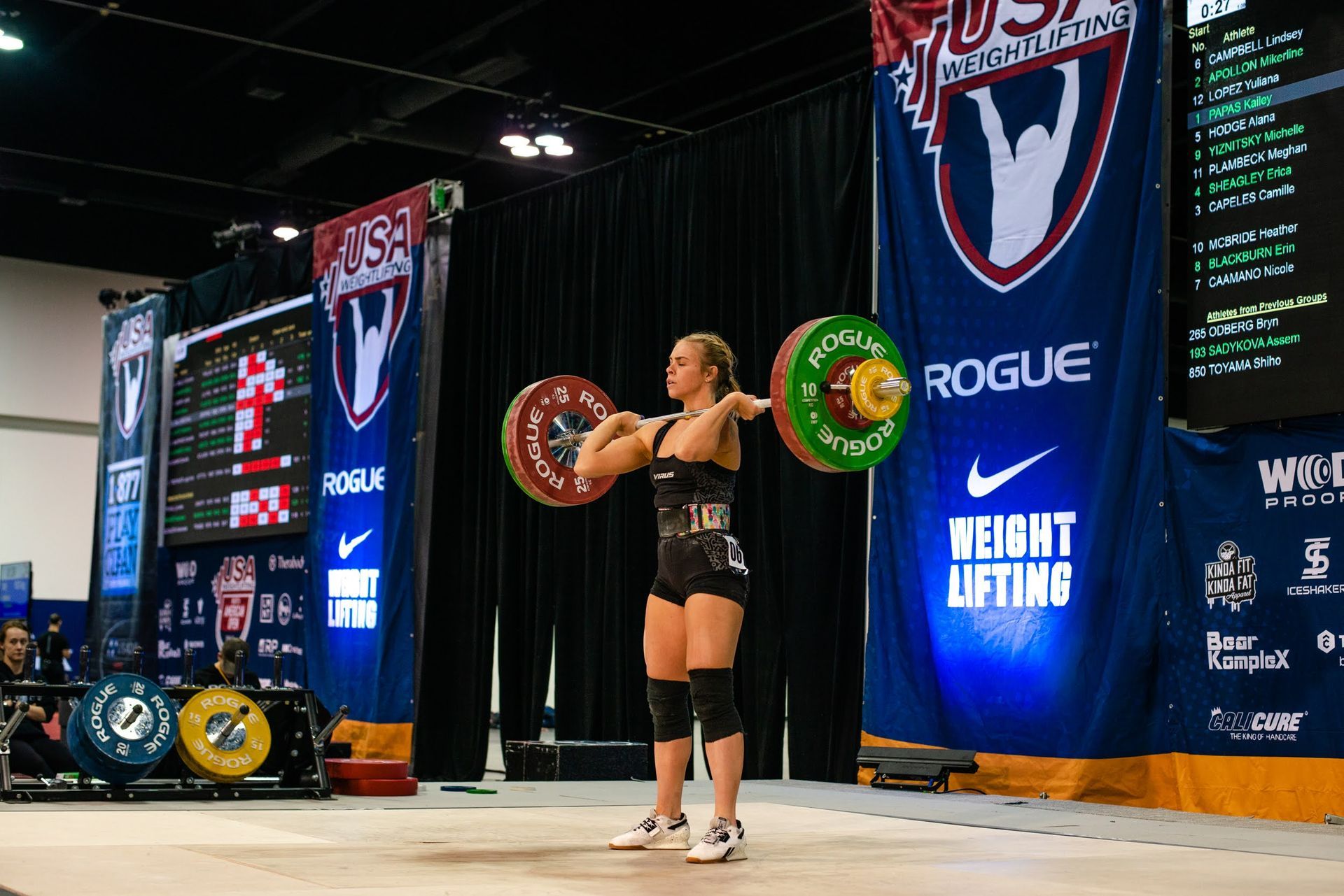 A female weightlifter holding a barbell overhead during a competition. USA Weightlifting banner in the background.